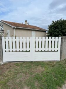 White wooden double gate with vertical slats in front of a beige house with a brown-tiled roof and a small yard behind.