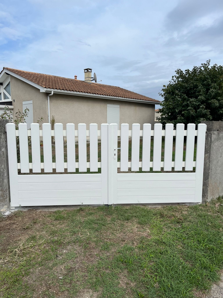 IMG-20260903-WA0003 | Menuiseries Hauxoises | Menuiseries Hauxoises White wooden double gate with vertical slats in front of a beige house with a brown-tiled roof and a small yard behind.